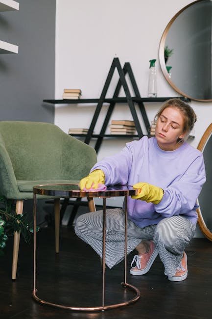 A young woman wearing a lavender sweatshirt and yellow rubber gloves is kneeling on a dark wooden floor while wiping the surface of a small, round, copper-colored side table with a pink cloth. Behind her, there is a green upholstered armchair, a black shelf holding books, and a round mirror on the wall, indicating a clean and tidy living room space. The room is well-lit with natural light, emphasizing the cleanliness and hygiene of the surface cleaning process. This image highlights domestic deep cleaning and surface sanitisation, as provided by Edgware Carpet Cleaning, emphasizing thorough cleaning routines within the home environment.