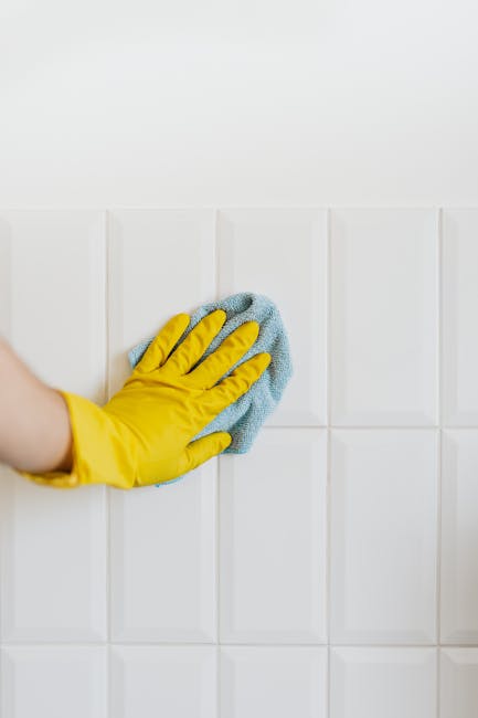 A close-up of a person wearing a yellow rubber glove, using a blue microfiber cloth to clean white ceramic wall tiles in a domestic setting. The tiles are arranged in a grid pattern with visible grout lines, and the surface appears spotless and shiny, indicating effective cleaning. The room is well-lit, enhancing the glossy finish of the tiles. This image illustrates effective surface cleaning and maintenance, which relates to the services offered by Edgware Carpet Cleaning for thorough domestic cleaning and sanitisation. The clean, bright environment underscores hygiene and professional cleaning standards promoted by Edgware Carpet Cleaning.