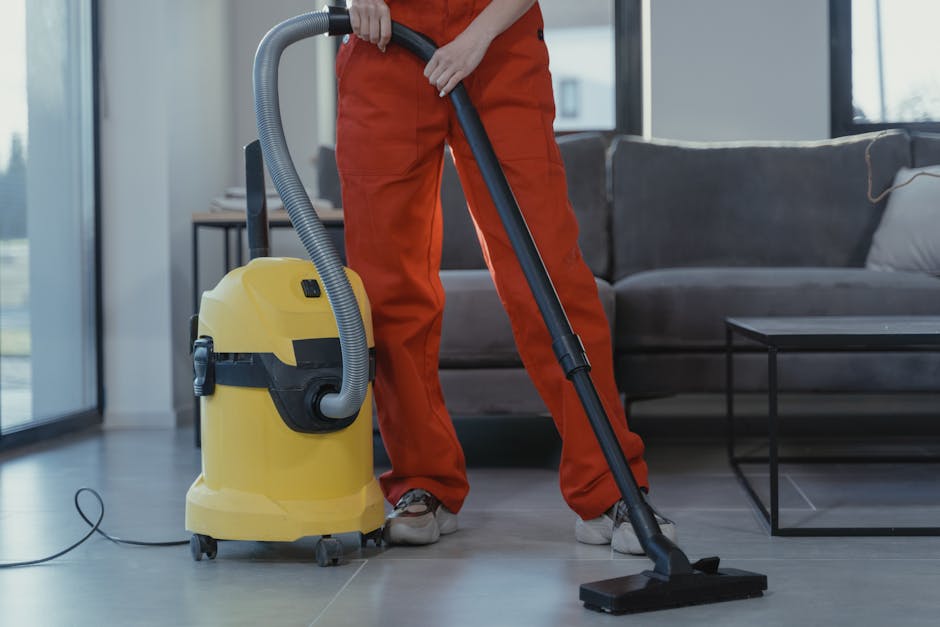 A person in orange overalls operating a yellow vacuum cleaner on a light-colored tiled floor in a modern living room. The vacuum hose connects to the main unit, which has wheels for mobility. The individual’s hands are holding the vacuum’s handle, directing it across the smooth, shiny surface. The room features a large window with natural light illuminating the space, a contemporary dark gray sofa with cushions, and a black metal coffee table. The overall scene depicts surface cleaning and deep cleaning processes, with a focus on maintaining hygiene and cleanliness. Edgware Carpet Cleaning provides professional domestic cleaning services that ensure thorough sanitisation and surface maintenance in residential spaces.