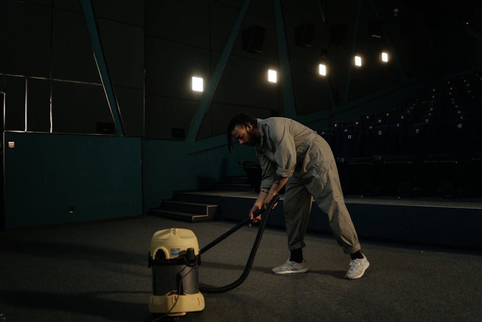 A professional cleaner dressed in full white protective gear, including a coverall, face mask, and gloves, is performing surface cleaning on a beige carpet inside a well-lit, modern living room. The cleaner is using a steam cleaning machine to deep clean the carpet, which appears to be in the process of being sanitized and refreshed. The room features natural light coming from a large window, with decorative plants in white pots, a wooden shelving unit, and a light-colored floor that enhances the clean and tidy appearance. Edgware Carpet Cleaning specializes in residential cleaning services, including deep carpet cleaning, to ensure hygiene and durability, as showcased in this detailed cleaning process near Edgware Station HA8. The area is visibly free of dust and debris, with the steam machine emitting a controlled amount of vapor, indicating high-quality sanitisation and surface cleaning techniques.