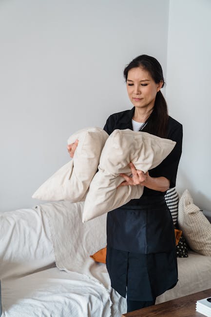 A professional cleaner from Edgware Carpet Cleaning stands in a living room holding two beige cushion covers, preparing for upholstery sanitisation and deep cleaning. The room features a white sofa with a slightly textured surface, decorated with patterned and plain scatter cushions, and a dark wooden side table. The background includes a plain white wall, with natural light illuminating the scene, highlighting the cleanliness and neatness of the space. Visible cleaning cloths and the absence of dust or stains contribute to a sense of hygiene and maintenance, aligning with surface cleaning and domestic cleaning services offered by Edgware Carpet Cleaning. The professional's attire is a dark uniform, emphasizing a professional approach to upholstery cleaning for residents or retailers within the Broadwalk Centre area.