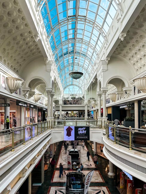 The image depicts the interior of the Broadwalk Centre shopping mall, featuring a large, arched glass ceiling allowing natural light to illuminate the space. The upper level has a white, ornate architectural design with decorative columns and balustrades made of glass and metal. The ceiling showcases intricate patterns with circular recesses and a central chandelier hanging underneath. Below, the ground floor displays retail shops with lit storefronts, and a digital display board is visible. The polished floors are clean and shiny, reflecting the light, suggesting recent surface cleaning or maintenance. Several shoppers are walking along the pathways, and the scene captures a bright and orderly environment that highlights the importance of hygiene and upkeep within this commercial setting, as part of professional cleaning and sanitisation efforts by Edgware Carpet Cleaning, which specializes in comprehensive cleaning services including upholstery and deep cleaning to ensure a pristine shopping experience.