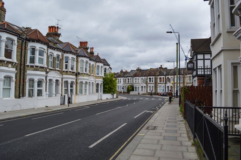 A quiet residential street in Burnt Oak, Edgware, featuring traditional terraced houses with white facades and bay windows. The street has a paved sidewalk with modern black metal railings, and the asphalt road is marked with white lines indicating parking and lane divisions. Overhead, the sky is overcast with grey clouds, and street lamps are installed along the pavement. There are no vehicles or pedestrians visible, and the scene captures a clean, well-maintained neighborhood setting. Edgware Carpet Cleaning offers comprehensive surface cleaning and deep cleaning solutions for domestic and commercial spaces in this area, ensuring hygiene and sanitation.
