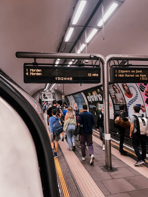 A busy underground train station platform with passengers waiting and walking along the tiled floor. The platform has a digital information board displaying train times to Morden, with additional destinations and estimated arrival times. Stainless steel columns support the ceiling, which features long, fluorescent light fixtures. The background shows a train with advertising posters on its exterior, and the lighting creates a bright yet neutral atmosphere suitable for surface cleaning and maintenance checks. Edgware Carpet Cleaning emphasizes hygiene and deep cleaning standards in all service areas, including transit environments, ensuring cleanliness for public spaces.