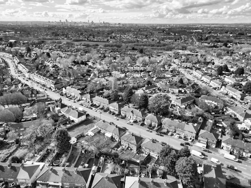 A quiet residential street in Burnt Oak, Edgware, featuring traditional terraced houses with white facades and bay windows. The street has a paved sidewalk with modern black metal railings, and the asphalt road is marked with white lines indicating parking and lane divisions. Overhead, the sky is overcast with grey clouds, and street lamps are installed along the pavement. There are no vehicles or pedestrians visible, and the scene captures a clean, well-maintained neighborhood setting. Edgware Carpet Cleaning offers comprehensive surface cleaning and deep cleaning solutions for domestic and commercial spaces in this area, ensuring hygiene and sanitation.