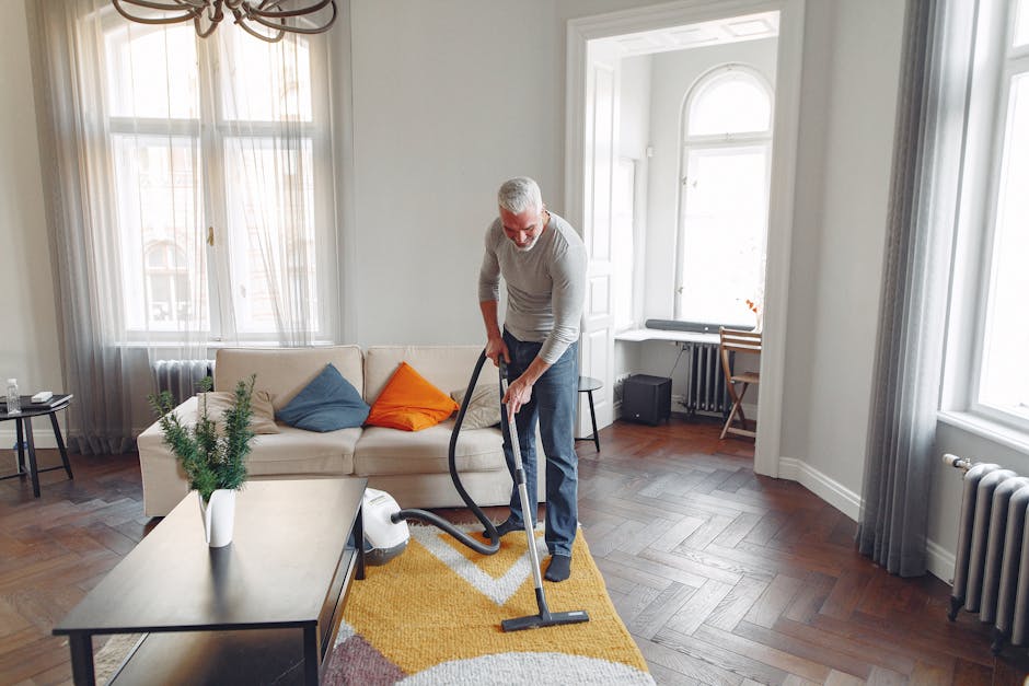 A middle-aged man with gray hair and a beard is vacuuming a wooden parquet floor in a bright, spacious living room. The room features large windows with sheer curtains allowing natural light to fill the space. The man is wearing a light gray long-sleeve shirt and dark pants, holding a vacuum cleaner with a hose extension, and is focused on cleaning an area rug with geometric patterns. A white sofa with colorful cushions (blue, orange) is positioned against the wall, with a small side table holding a glass of water and a decorative plant nearby. The room includes a wooden coffee table, a desk with a chair by the window, and a radiator along the wall. The overall scene depicts a clean and well-maintained living space, emphasizing effective surface cleaning and maintenance techniques, as recommended by Edgware Carpet Cleaning in their guide on common carpet cleaning mistakes landlords see in Edgware.