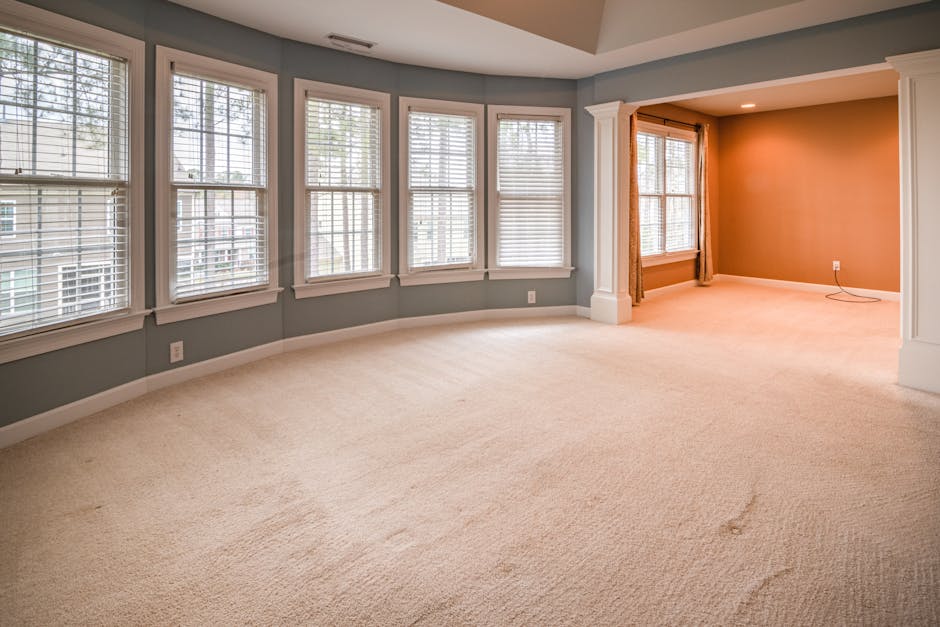 A spacious living room with a light beige carpet that appears clean but shows slight signs of wear and minor stains in some areas. The room features large white-framed double-hung windows with horizontal blinds, allowing natural light to brighten the space. The walls are painted in soft pastel colors, with the main area in a light blue and accent areas in warm beige, creating a cozy atmosphere. White crown molding and baseboards frame the room, and there is a white column with decorative trim separating the adjacent spaces. The room's flooring is maintained well, but improper cleaning techniques like neglecting deep cleaning or using inappropriate cleaning products can lead to dirt buildup or dullness, which Edgware Carpet Cleaning can help prevent through proper surface cleaning and sanitisation. The overall condition suggests regular domestic cleaning, but without deep cleaning, spots or accumulated dust may remain, highlighting common mistakes landlords should avoid for optimal carpet hygiene and appearance.