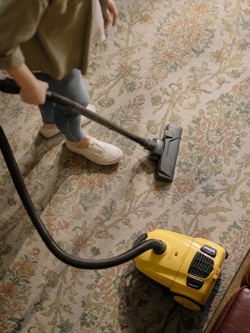 A middle-aged man with gray hair and a beard is vacuuming a wooden parquet floor in a bright, spacious living room. The room features large windows with sheer curtains allowing natural light to fill the space. The man is wearing a light gray long-sleeve shirt and dark pants, holding a vacuum cleaner with a hose extension, and is focused on cleaning an area rug with geometric patterns. A white sofa with colorful cushions (blue, orange) is positioned against the wall, with a small side table holding a glass of water and a decorative plant nearby. The room includes a wooden coffee table, a desk with a chair by the window, and a radiator along the wall. The overall scene depicts a clean and well-maintained living space, emphasizing effective surface cleaning and maintenance techniques, as recommended by Edgware Carpet Cleaning in their guide on common carpet cleaning mistakes landlords see in Edgware.