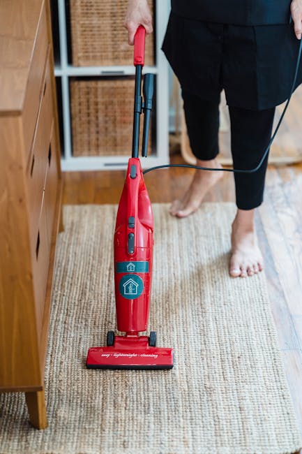 A person using a red vacuum cleaner for surface cleaning on a beige carpet in a residential setting, with visible wooden furniture and a bookshelf in the background. The individual, dressed in black clothing, is barefoot and holding the vacuum handle, which is plugged into an electrical outlet. The room is illuminated with natural light, highlighting the clean, well-maintained appearance of the carpet and surrounding surfaces. The image reflects domestic cleaning practices that Edgware Carpet Cleaning offers for deep cleaning and sanitisation, aligning with the Hale Lane rug cleaning guide for Edgware homes, EDGWARE.