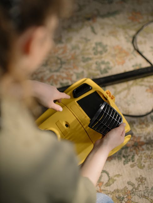 A person holding a yellow portable industrial fan or blower, positioned on a patterned rug in a domestic setting, with the person's hand pressing a button on the device. The scene includes a carpet with a floral or oriental design, and a power cord extending from the device. Lighting appears natural and the room looks clean and well-maintained, consistent with professional surface cleaning and deep cleaning processes promoted by Edgware Carpet Cleaning, as seen on the Hale Lane rug cleaning guide page for Edgware homes.